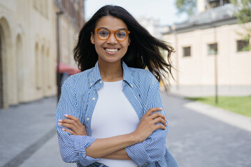 Fototapeta premium Portrait of beautiful smiling African American woman looking at camera, standing on the street. Young confident businesswoman with arms crossed wearing stylish eyeglasses posing for pictures outdoors
