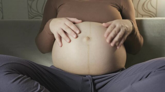 young pregnant woman stroking big belly on couch, making heart shape with hands