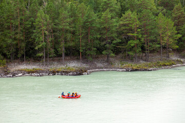Fototapeta na wymiar A team of people rafting in orange life jackets on an rubber boat of blue and yellow colors along a mountain river against the background of a rocky shore with a forest.