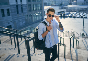 young man with a black backpack standing on a city street holding sunglasses in his hands.