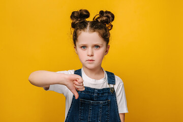 Portrait of serious little girl look at camera show thumb down gesture symbol of negative feedback and dislike, isolated over yellow studio background wall. Disagreement and rejection concept © Yura Yarema