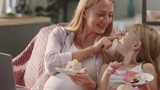 Cheerful pregnant mother booping nose of cute little daughter and laughing while sitting together on sofa at home and enjoying sweet cakes