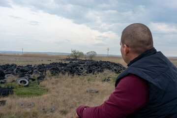 The man sits thoughtfully and looks at a huge pile of car tires. The problem of ecology and waste...