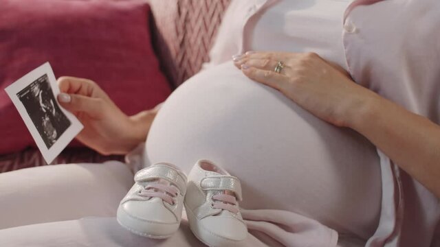 Close up view of expectant mother sitting on sofa with cute bootees on lap, holding fetal ultrasound image and rubbing pregnant belly while waiting for baby