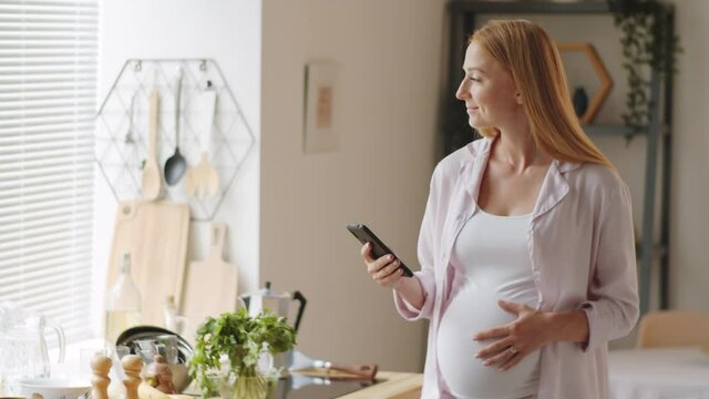 Medium shot of beautiful expectant mother standing at home, touching her pregnant belly and browsing the mobile Internet on smartphone