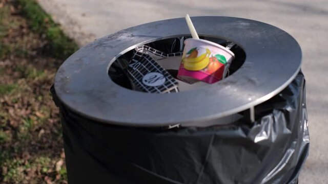 Overflowing garbage bin on the street in and out of focus. Colorful take away packagings, ice cream cup with a plastic spoon and a 28 cm pizza box in a black dustbin. Garbage concept, sunny day, 4k.