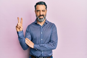 Middle age man with beard and grey hair wearing business clothes smiling with happy face winking at the camera doing victory sign. number two.