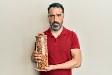 Fototapeta premium Middle age man with beard and grey hair holding jar with macaroni pasta clueless and confused expression. doubt concept.