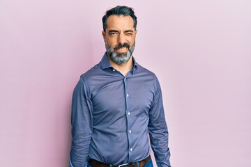 Middle age man with beard and grey hair wearing business clothes winking looking at the camera with...