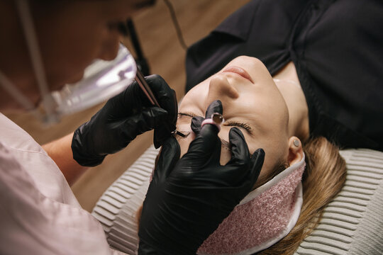 eyebrow microblading. A master in a transparent mask on his face and black gloves holds a handpiece with a shading needle over the eyebrow of the model.