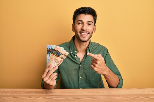Young handsome man holding canadian dollars cheerful with a smile on face pointing with hand and finger up to the side with happy and natural expression