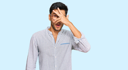 Young handsome man wearing casual clothes and glasses peeking in shock covering face and eyes with hand, looking through fingers with embarrassed expression.