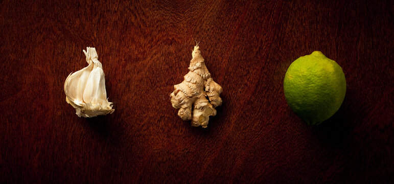 A detail arrangement of garlic, ginger and lime, side by side on a dark wooden table.    