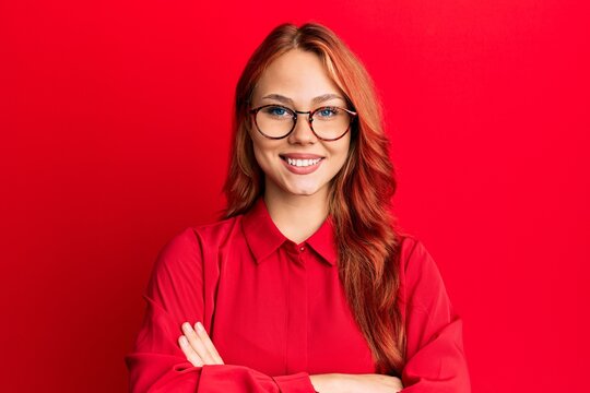 Young beautiful redhead woman wearing casual clothes and glasses over red background happy face smiling with crossed arms looking at the camera. positive person.