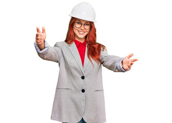Young redhead woman wearing architect hardhat looking at the camera smiling with open arms for hug. cheerful expression embracing happiness.