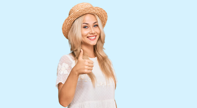 Young blonde girl wearing summer hat doing happy thumbs up gesture with hand. approving expression looking at the camera showing success.