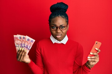 Young african american woman using smartphone holding south africa rand banknotes clueless and...