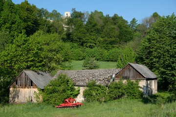 Obraz premium Rural landscape with farm and red disc cultivator between green trees at foot of hill with Pilcza castle, Krakow-Czestochowa Upland, Silesia, Poland