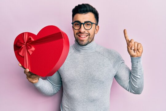 Young hispanic man holding valentine gift smiling with an idea or question pointing finger with happy face, number one