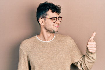 Young hispanic man wearing casual clothes and glasses looking proud, smiling doing thumbs up gesture to the side