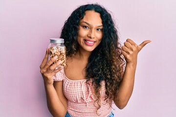 Young latin woman holding bowl with walnuts pointing thumb up to the side smiling happy with open mouth