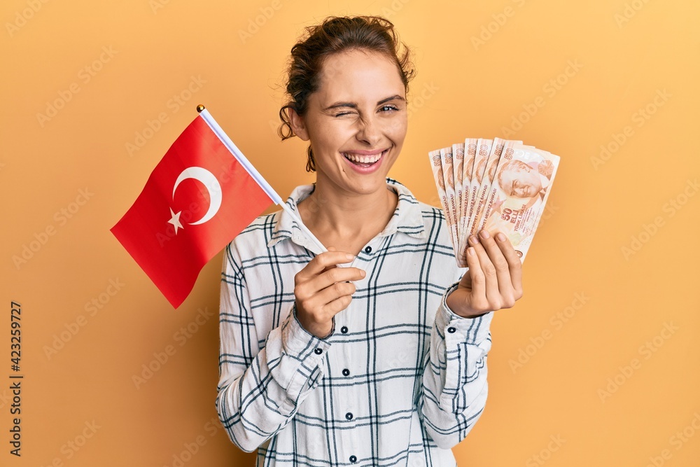 Poster Young brunette woman holding turkey flag and liras banknotes winking looking at the camera with sexy expression, cheerful and happy face.