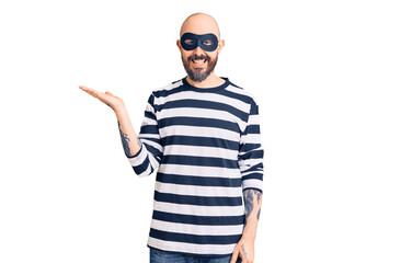 Young handsome man wearing burglar mask smiling cheerful presenting and pointing with palm of hand looking at the camera.