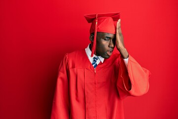 Young african american man wearing graduation cap and ceremony robe surprised with hand on head for mistake, remember error. forgot, bad memory concept.