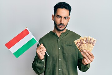 Young hispanic man holding hungary flag and forints relaxed with serious expression on face. simple...
