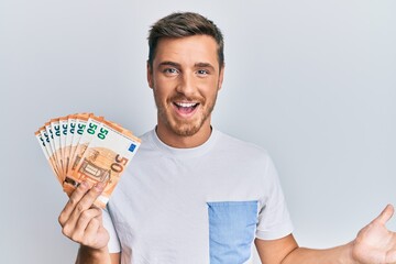 Handsome caucasian man holding 50 euro banknotes celebrating achievement with happy smile and...
