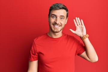 Handsome caucasian man wearing casual red tshirt waiving saying hello happy and smiling, friendly welcome gesture