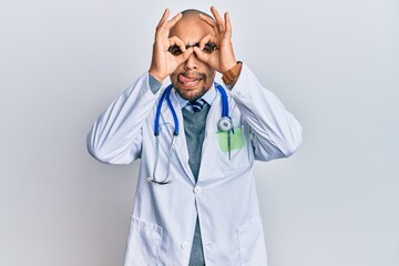 Hispanic adult man wearing doctor uniform and stethoscope doing ok gesture like binoculars sticking tongue out, eyes looking through fingers. crazy expression.