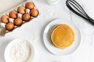 composition of small desserts for baking. eggs, muffins, flour, milk, whisk and spoon on a light background. Close-up of baked pancakes on a white plate.