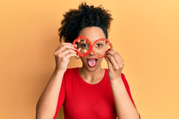 Young african american girl holding red pepper as a glasses sticking tongue out happy with funny expression.