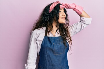 Young brunette woman with curly hair wearing cleaner apron and gloves very happy and smiling looking far away with hand over head. searching concept. - obrazy, fototapety, plakaty