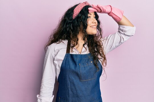 Young brunette woman with curly hair wearing cleaner apron and gloves very happy and smiling looking far away with hand over head. searching concept.