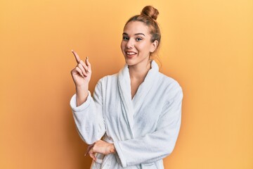 Young blonde woman wearing bathrobe with a big smile on face, pointing with hand and finger to the side looking at the camera.