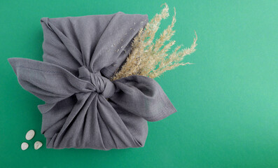 A gift wrapped in a gray linen napkin with branches of pampas grass on a green background with...