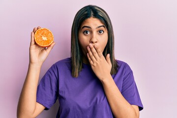 Young latin woman holding fresh orange slice covering mouth with hand, shocked and afraid for...