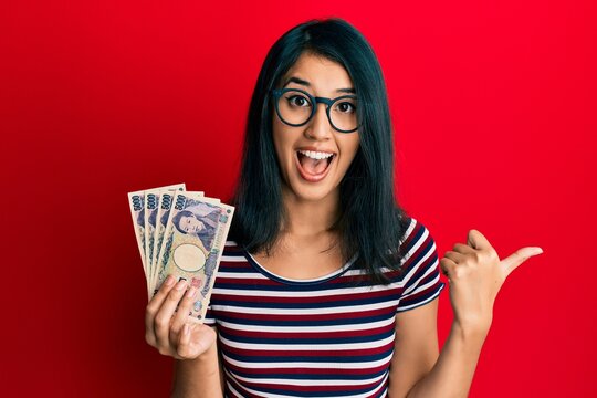 Beautiful asian young woman holding 5000 japanese yen banknotes pointing thumb up to the side smiling happy with open mouth