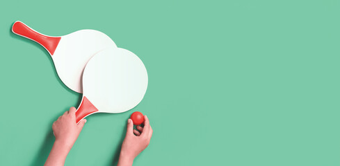 Hands holding one of two white and red beach tennis paddle racket with red ping pong ball against green background. Flat lay with copy space. Outdoors summer activity, education break kids fun