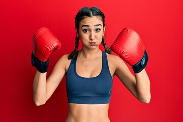 Young brunette girl using boxing gloves wearing braids puffing cheeks with funny face. mouth inflated with air, catching air.