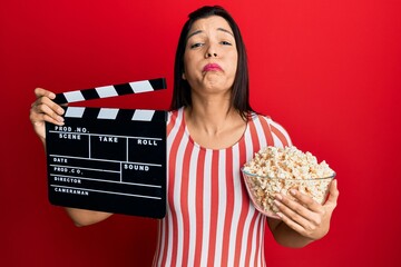 Young latin woman holding video film clapboard and popcorn puffing cheeks with funny face. mouth...