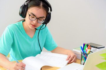 Asian woman student teenage girl with glasses headphones sitting happy smile looking writing notes at book laptop computer on table learning online study education from the class of university at home