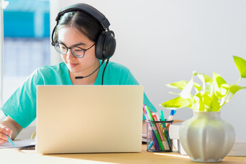 Asian woman student teenage girl with glasses headphones sitting happy smile looking writing notes at book laptop computer on table learning online study education from the class of university at home