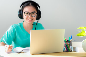 Portrait young Asian female student with headphones glasses smile looking up a note on the book a teenage girl is happy to university internet distance study online class on a laptop computer at home