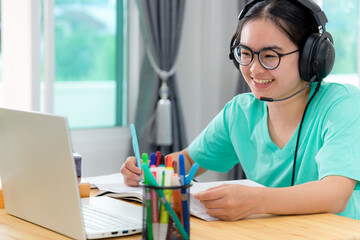 Asian young woman student with glasses headphones girl study happy writing note on a book looking video conference laptop computer university class online internet learning distance education at home