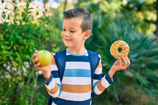 Adorable caucasian student boy smiling happy holding donut and bottle of water at the park.