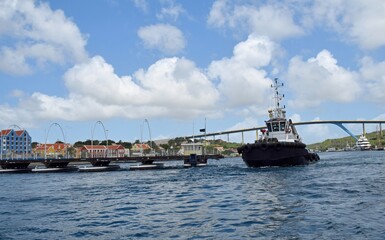 Fototapeta premium Pilot tug boat passing Queen Emma Bridge pontoon bridge across St. Anna Bay in Willemstad Curaçao; partially open with the colorful buildings of Otrobanda and the Queen Juliana Bridge in the backgroun