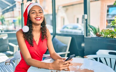 Young african american girl wearing christmas hat using smartphone sitting on the table at terrace.